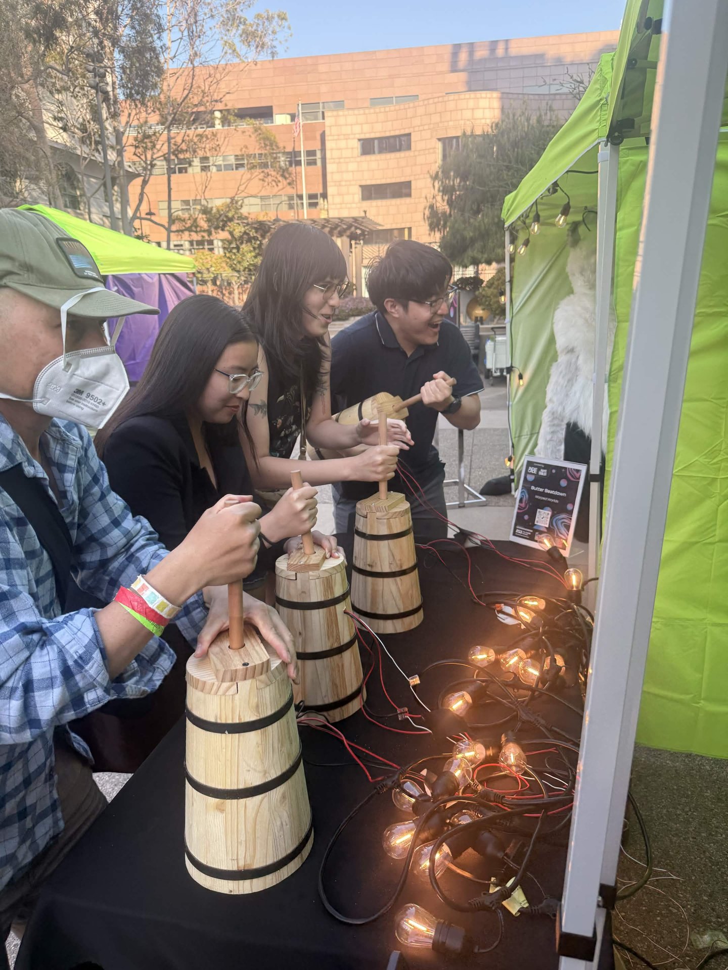 Players lined up at a table using wooden Butter Beatdown churn controllers at IndieCade